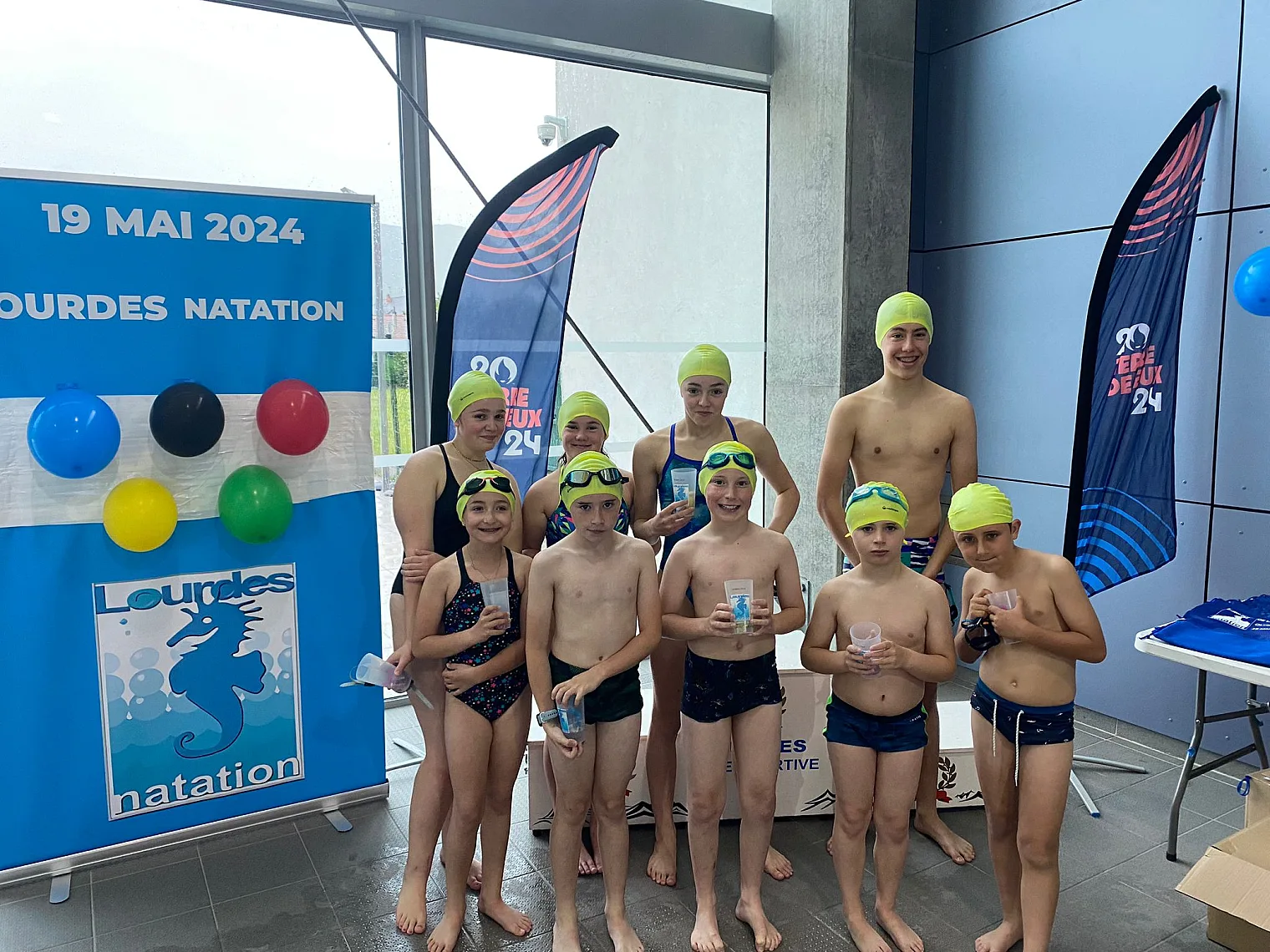Jeunes nageurs du club en bonnet de bain devant les oriflammes Lourdes Natation et Terre de Jeux 2024